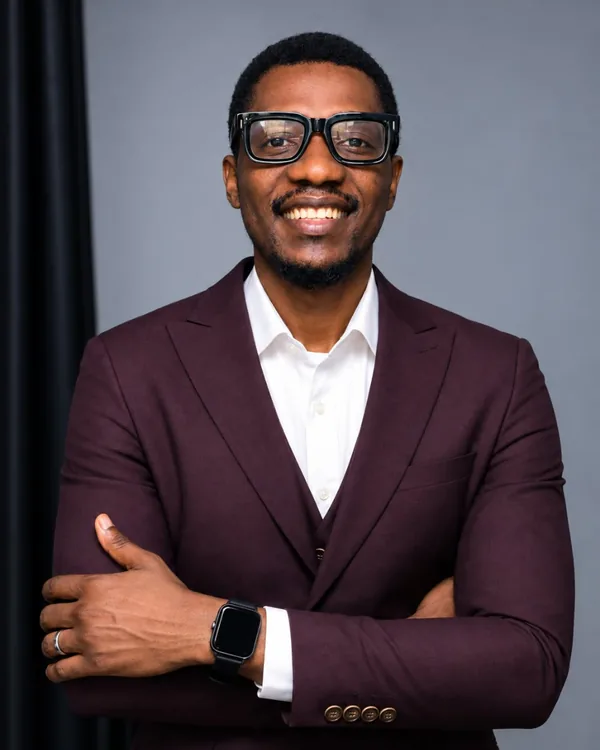 Paul Olatoye wearing glasses, a light blue shirt and a patterned tie, arms folded